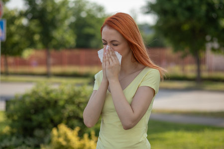 Young redhair woman sneezing in summer in front of blooming tree. Pollen Allergy symptomsの写真素材