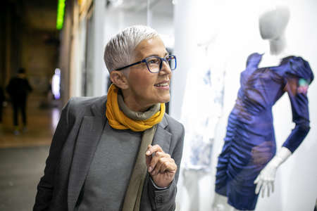 Smiling mature senior woman with short gray hair and eyeglasses looking in the shop window, city night sceneの写真素材
