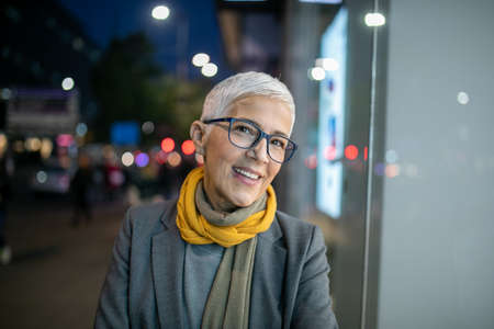 Smiling mature senior woman with short gray hair and eyeglasses walking on street, city night sceneの写真素材