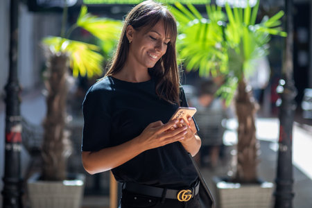 Beautiful smiling woman in black shirt texting on smartphone on street in city center. Palm in backgroundの写真素材