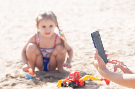 hands of a woman photographing a small three-year-old girl on her phone playing with a machine on...