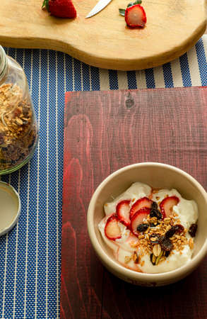 A deep plate with yogurt, granola, and strawberry, on a wooden table. Preparation of a light breakfast, yogurt, granola and strawberry ready to eat, next to the ingredients on a wooden tableの写真素材