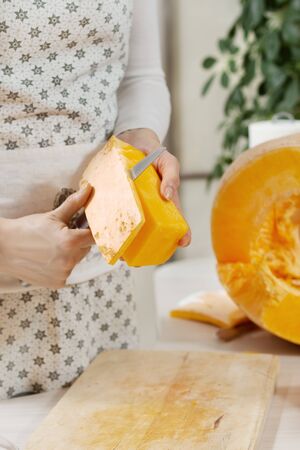 Female hands hold a piece of pumpkin and peel it off the skin with a kitchen knife. Half of the fresh large orange pumpkin lies on the kitchen counter.の写真素材