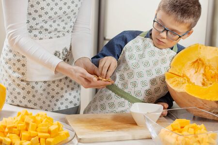 The woman and the boy in the kitchen aprons are standing at the counter. The boy takes from the hands of the woman fresh pumpkin seeds. The diced pumpkin lies on a glass plate.の写真素材