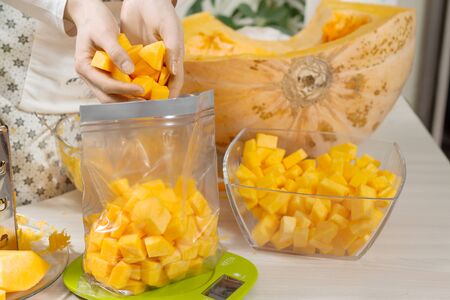 The woman puts diced fresh orange pumpkin in a plastic bag for storing food. The foil bag stands on the kitchen scale.の写真素材
