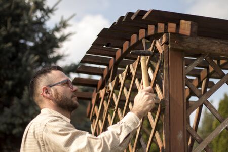 Man's hand holds a hammer and sticks a nail into a wooden garden pergola. Renovation and maintenance and repair of wooden garden furniture.の写真素材