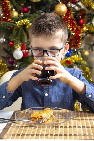 The boy is sitting at the table during dinner. He holds a glass with a drink in his hands and drinks from it. Christmas tree in the background.の写真素材