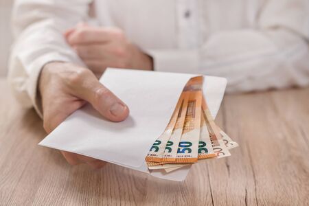 Man's hand holds a white envelope filled with Euro banknotes and hands it over the table.