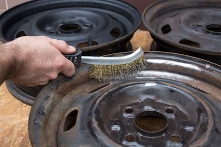 The mechanic cleans the steel wheel rim manually with a wire brush. Repair, regeneration and painting of automobile rims in a car workshop.の写真素材