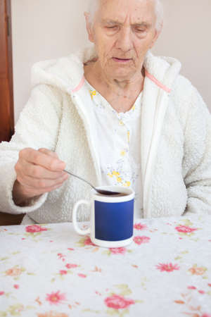 The gray-haired old man sits at the table and takes a cup of tea from a teaspoon.の写真素材