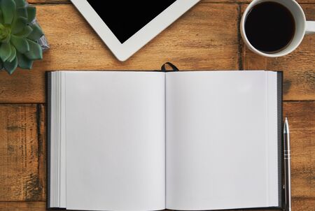 Blank page of open notebook with tablet and cup of coffee on wooden table desktop. Top view image.の写真素材