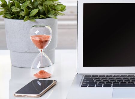 Close-up of sandglass with laptop and phone. Close-up of home office workspace, desktop.の写真素材