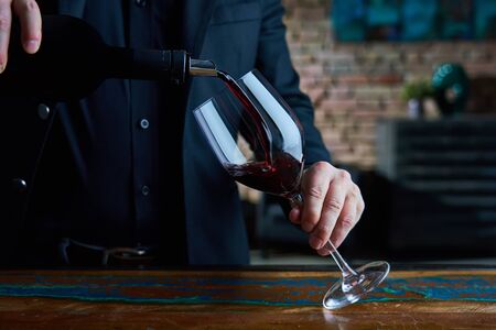 Man in black suit and black shirt pouring red wine into wine glass. Close up image of man holding...