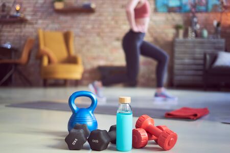 Woman exercising at home in living room. Focus on fitness equipments, barbell and kettlebell. Woman doing walking lunges exercise in the background. Concepts about home workout, fitness, sport and health.の写真素材