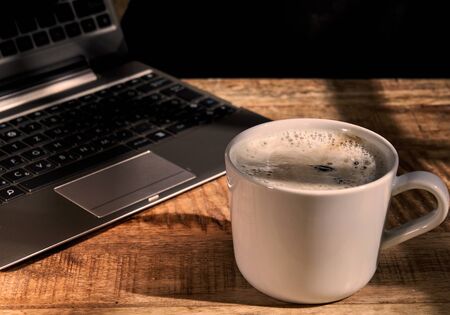 Close-up of cup of coffee with laptop next to it on wooden table.ï¿½の写真素材