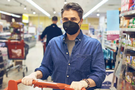 Man buying groceries in supermarket, wearing black medical face mask while shopping in grocery store. Covid-19 virus prevention.の写真素材