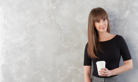 Indoor portrait of happy smiling woman drinking coffee, holding disposable paper cup, leaning...