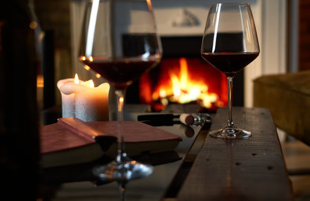 Two glasses of red wine with book and candle on table at home, fireplace in the background. Warm, dark colors.の写真素材