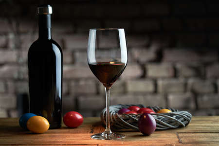 Easter decorations, colorful eggs, a glass of red wine with a bottle on table at home dark warm...