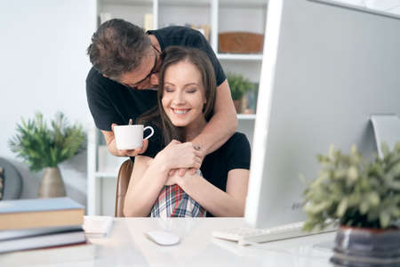 Happy loving couple drinking coffee at home in the morning, man hugging his girlfriend / wife at office desk.の写真素材