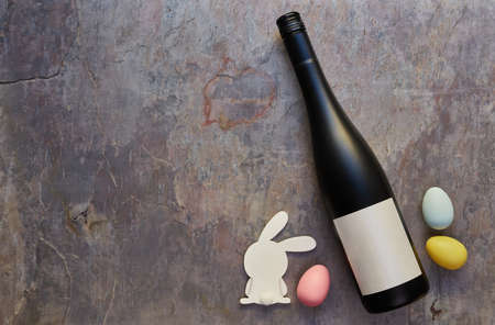 Top view of wine bottle with blank label and Easter eggs and white bunny on dark stone table...