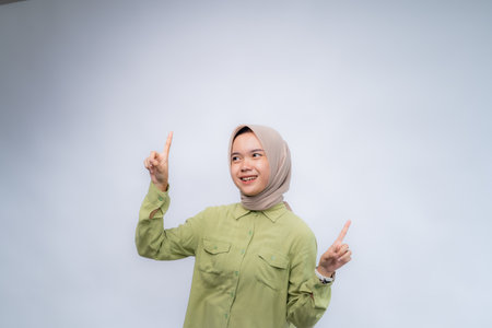 Look Up. Portrait of young asian woman wearing hijab Pointing Fingers Upward at Copy Space Advertising Your Text, Standing Isolated Over white Studio Backgroundの写真素材