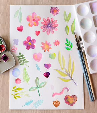 Closeup watercolor drawing on paper with brushes, palette and paints, different flowers, leaves, hearts paintedの写真素材
