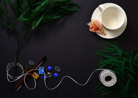 The background is black with a cup on a saucer, eucalyptus leaves, rope, watercolor paints, brushes....