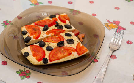 Omelet with tomatoes and olives. Two pieces on a dark glass plate, next to a table with a white...