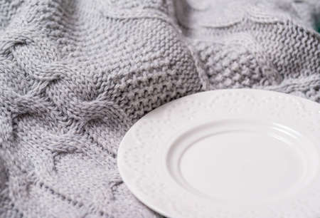 White plate on a gray knitted woolen blanket. Close-up of warm hand-knitted fabric, empty glossy ceramic plate in the corner.の写真素材