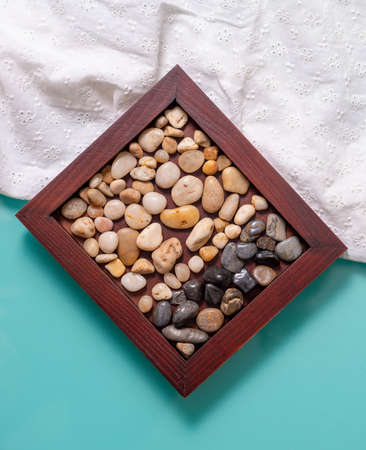 A wooden frame with two colors of sea pebbles on a turquoise table with a white cloth. The background is divided by a cloth in half, the frame is diagonal, empty space on the background for the text.の写真素材