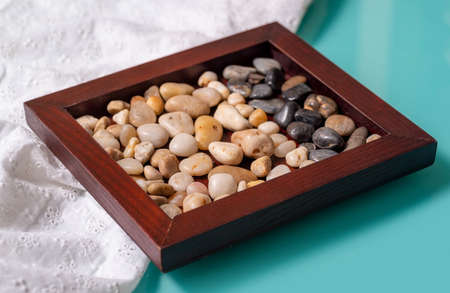 A wooden frame with two colors of sea pebbles on a turquoise table with a white cloth. The...