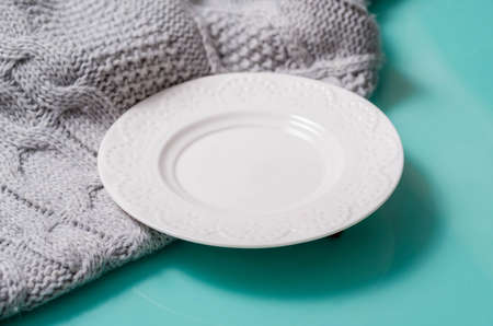The white plate stands on a turquoise glossy table and a gray woolen knitted fabric, a view from the side. The dish is empty, plaid large viscous warm and cozy, falls a shadow to the right.の写真素材