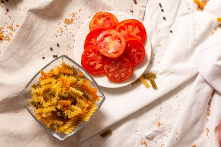 Top view of the ingredients for making pasta with vegetables and spices. Light background of mint tablecloth and towels, on which spices are scattered. Sliced tomatoes, pasta spirals.の写真素材