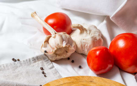 Three tomatoes and two heads of garlic on a white cloth. At the bottom is a fragment of a cutting board, several balls of allspice.の写真素材