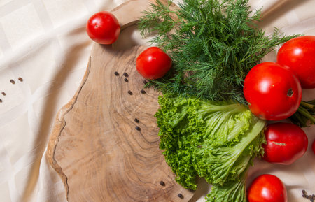Fragment of vegetables for salad on a wooden cutting board. Tomatoes, lettuce, dill lie partly on a beige tablecloth on the table, partly on a board. Peppercorns are scattered nearby. Top view.の写真素材