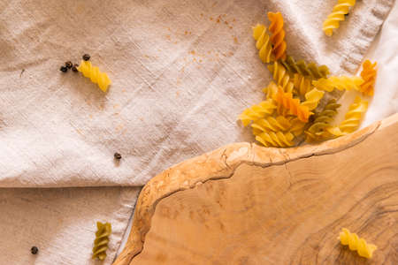 Fragment of cutting board curved shape of olive tree, scattered around pasta, garlic, pepper. They lie on a crud towel. Top view of the Italian cooking set. Horizontal photo macro.の写真素材