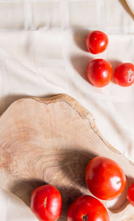 Fragment of cutting board made of olive tree, on which lie four tomatoes. The composition lies on the table with a tablecloth and other tomatoes. Folds on beige fabric in a large cage.の写真素材