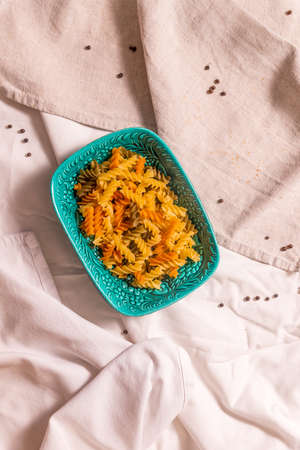 A turquoise ceramic bowl of colored pasta stands on a table covered with towels. View from above on...
