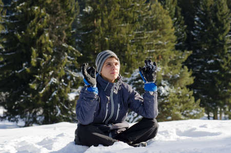 Teenager man sit on snow with winter jacket and gloves contemplating and think in yoga position with fir trees behindの写真素材