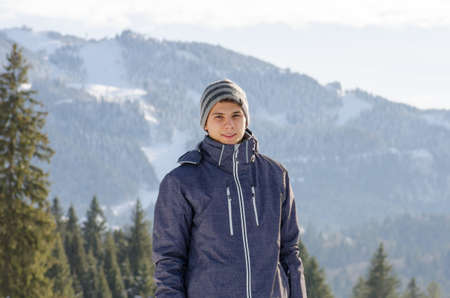 Teenager man smiling in winter jacket at mountains with ski slopes and fir trees in background