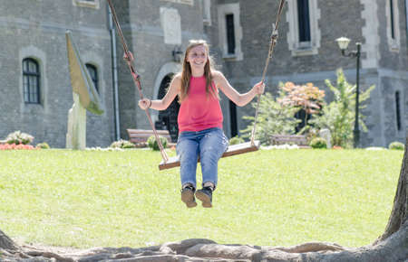 Young blonde woman smiling on wooden swing near old castle in nature