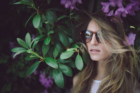 beautiful young girl in a white dress posing in a green bush with pink flowers, wearing sunglasses,...