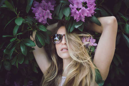 beautiful young girl in a white dress posing in a green bush with pink flowers, wearing sunglasses, looking into the camera, one eye hidden behind her hairの写真素材