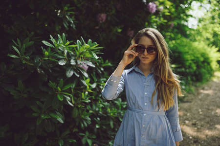 beautiful young girl in a blue dress walking through a park wearing sunglasses, bushes and flowers in the backgroundの写真素材