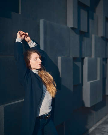 Beautiful blond woman leaning on a dark structured wall with her hands up and her eyes closedの写真素材