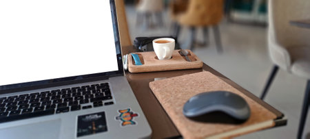 Workspace setting featuring laptop with blank screen, coffee, mouse, and notebook on table in cafe,...