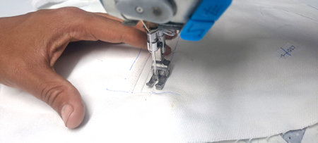 Close-up of tailor's hand guiding white fabric under sewing machine needle, creating a precise...