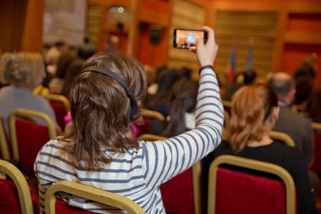 Man with headset at the conference . The audience wearing head phone for online translation . Unrecognizable people using in ear headphones for translation during event . Female video conference with phoneの写真素材