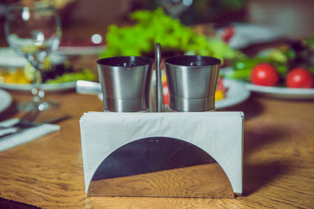 A table in a cafe or restaurant. White napkins in a metal napkin holder, spices salt and pepper on a wooden table, panoramic view . metal napkin holder isolated on white background.の写真素材